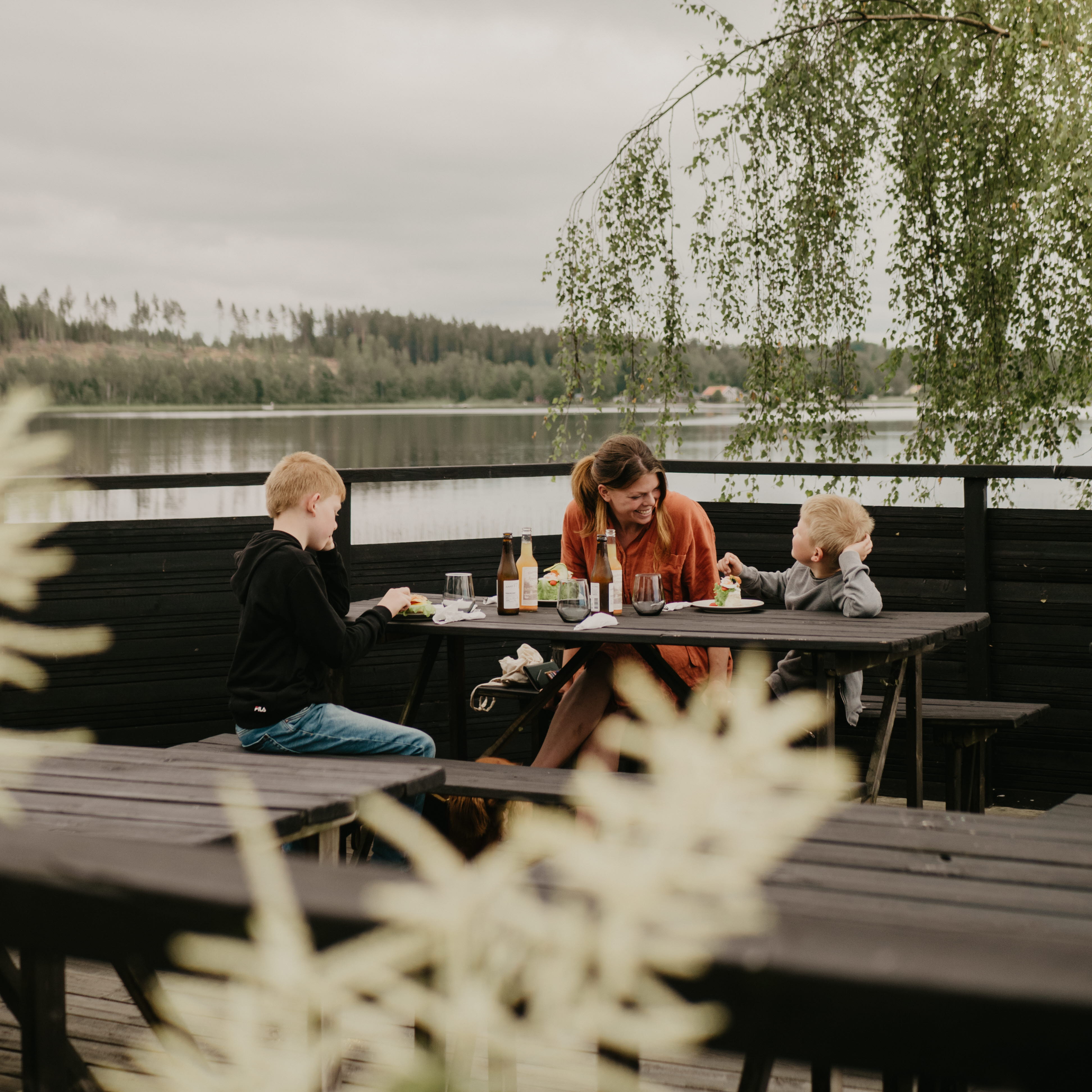 Mamma och två barn sitter och fikar på brygga vid vattnet