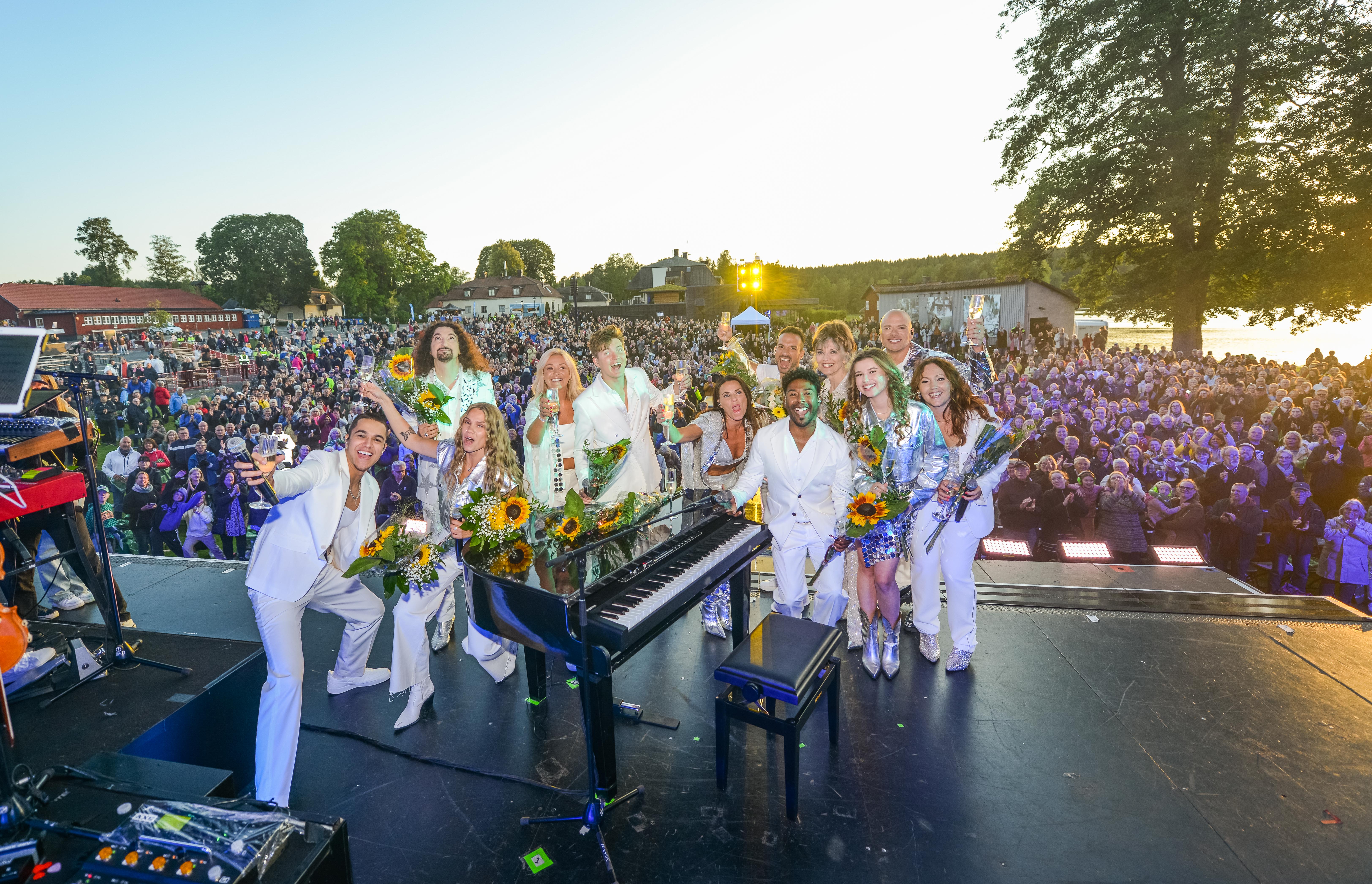Artister står vid ett piano på en scen. Stor publik i bakgrunden.