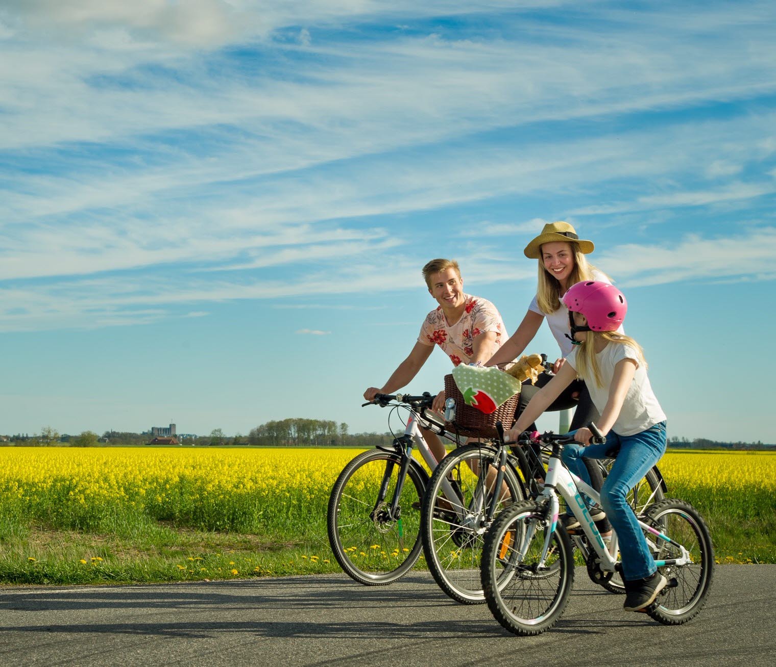 En familj är ute och cyklar på en landsväg en solig sommardag. I bakgrunden står stora gula fält.
