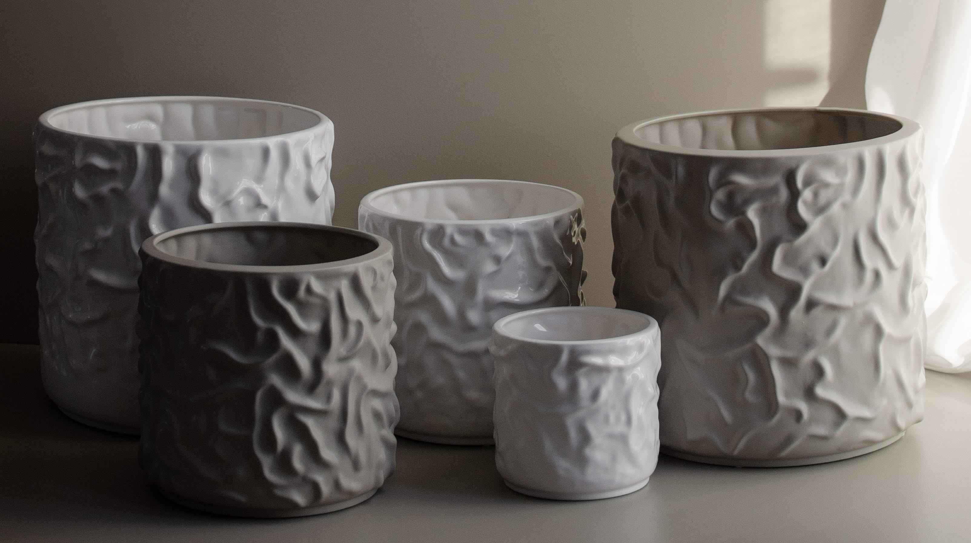 Fem krukor i olika storlekar i beige och vitt står framför en beige vägg. 