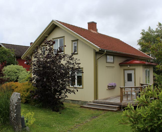 Ett ljust grön/gult litet hus i en lummig fin trädgård.