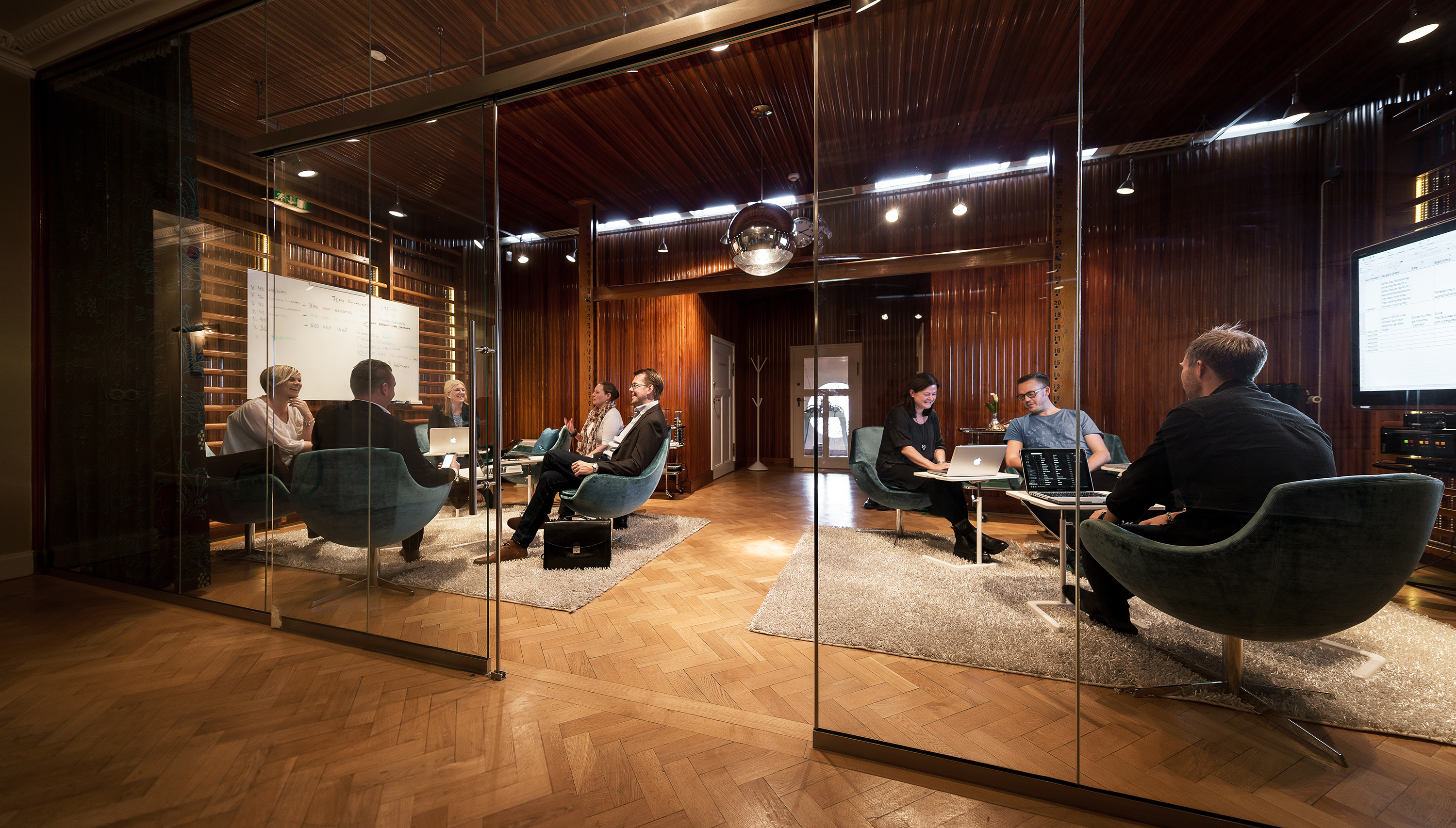 Behind a glass wall some people are sitting in some chairs with computers on the tables in front of them.