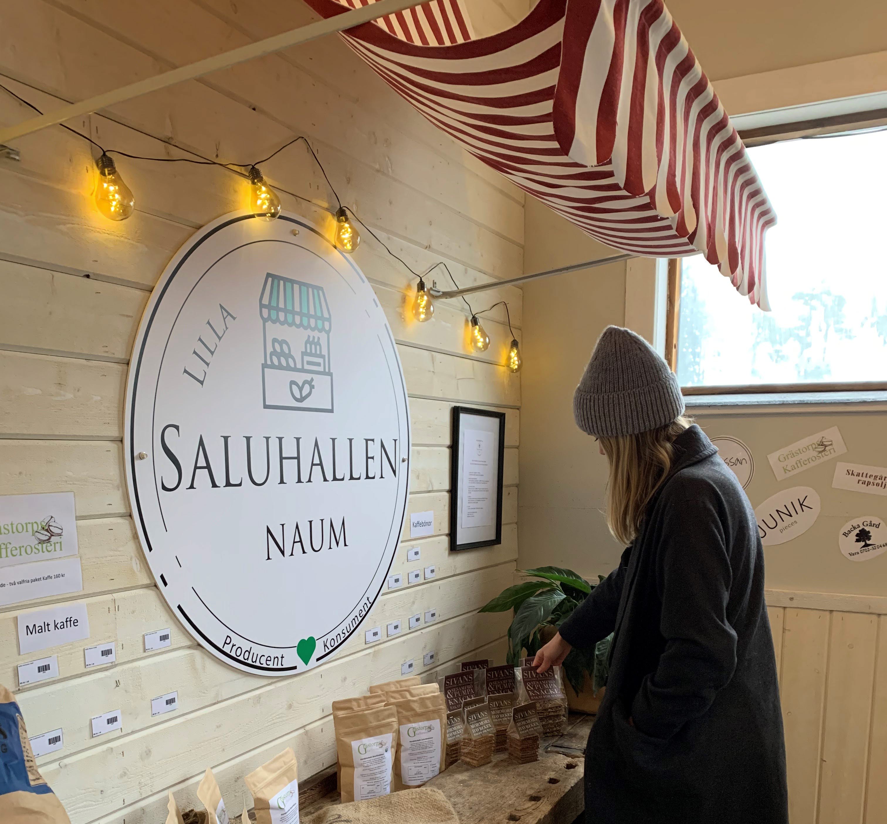 Kvinna i grå mössa och svart kappa som kollar i en butik. På väggen är det en skylt där det står Lilla Saluhallen Naum.