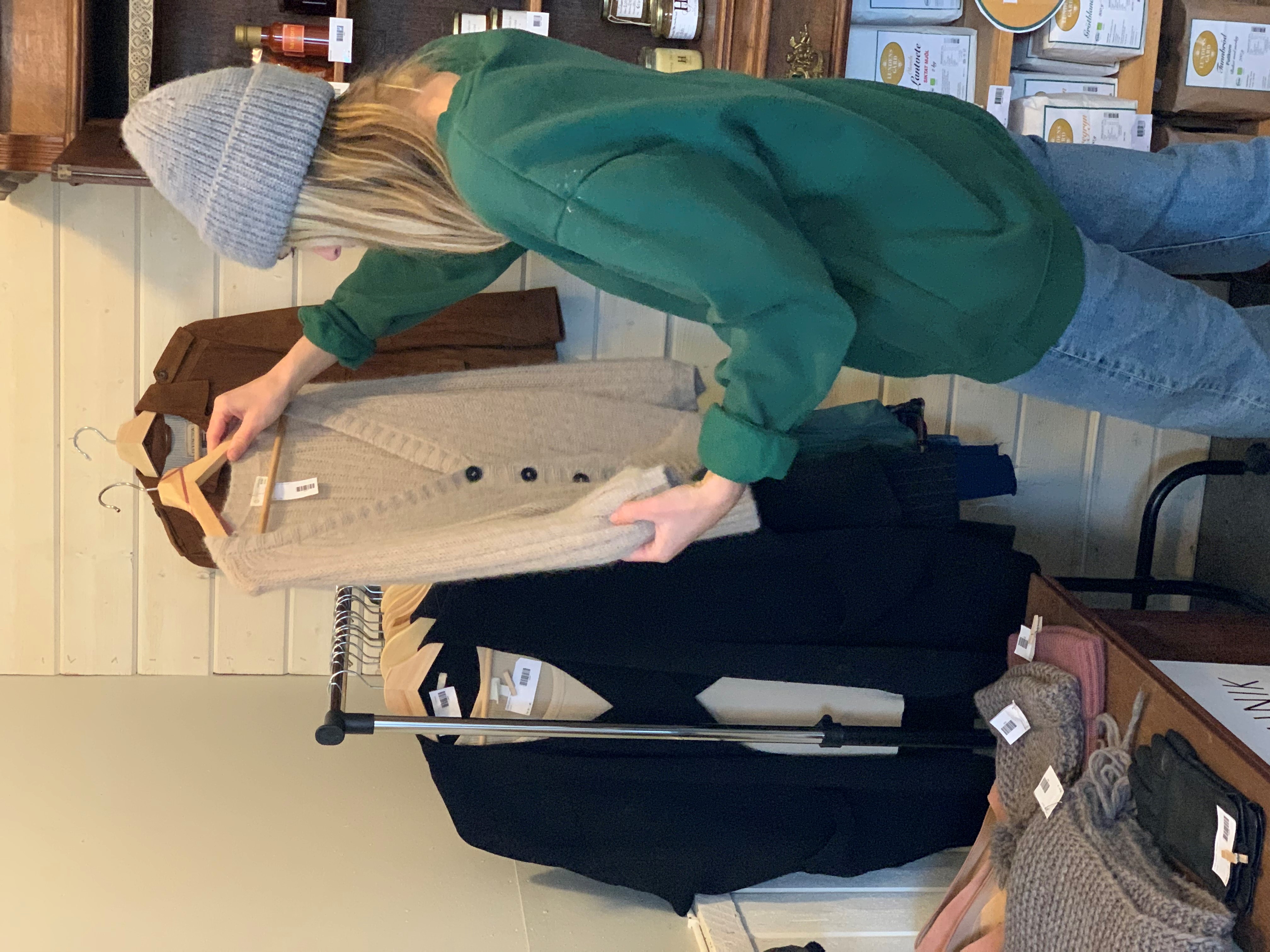 Kvinna i grön tröja och grå mössa kollar på en beige tröja. 