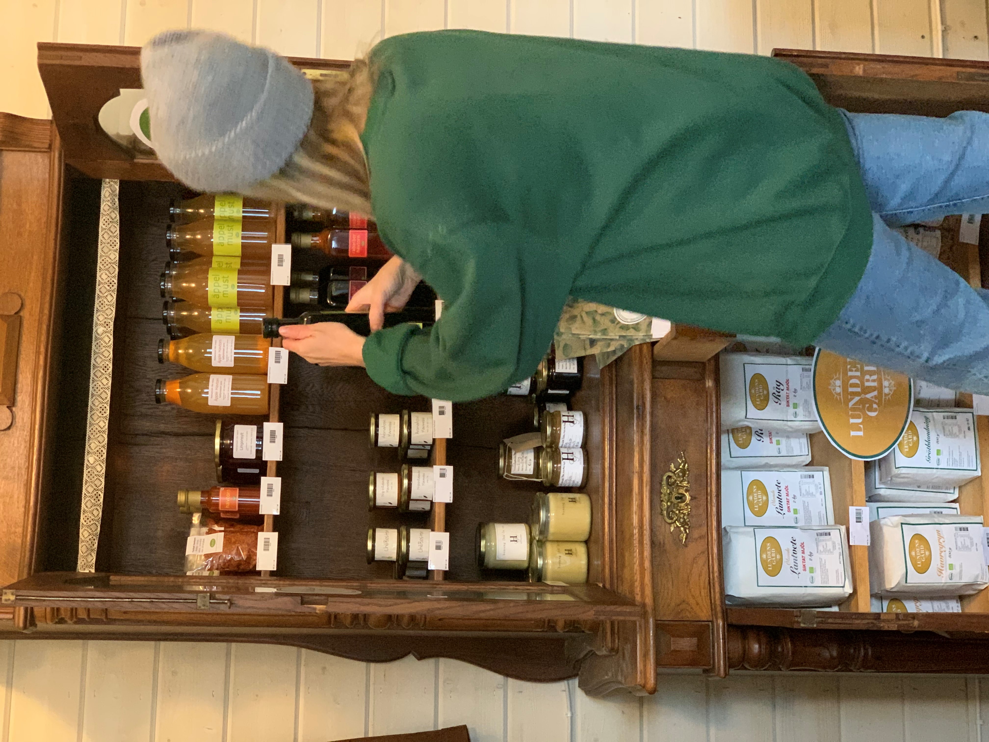 Kvinna i grön tröja och grå mössa kollar på en hylla med olika glasflaskor. 