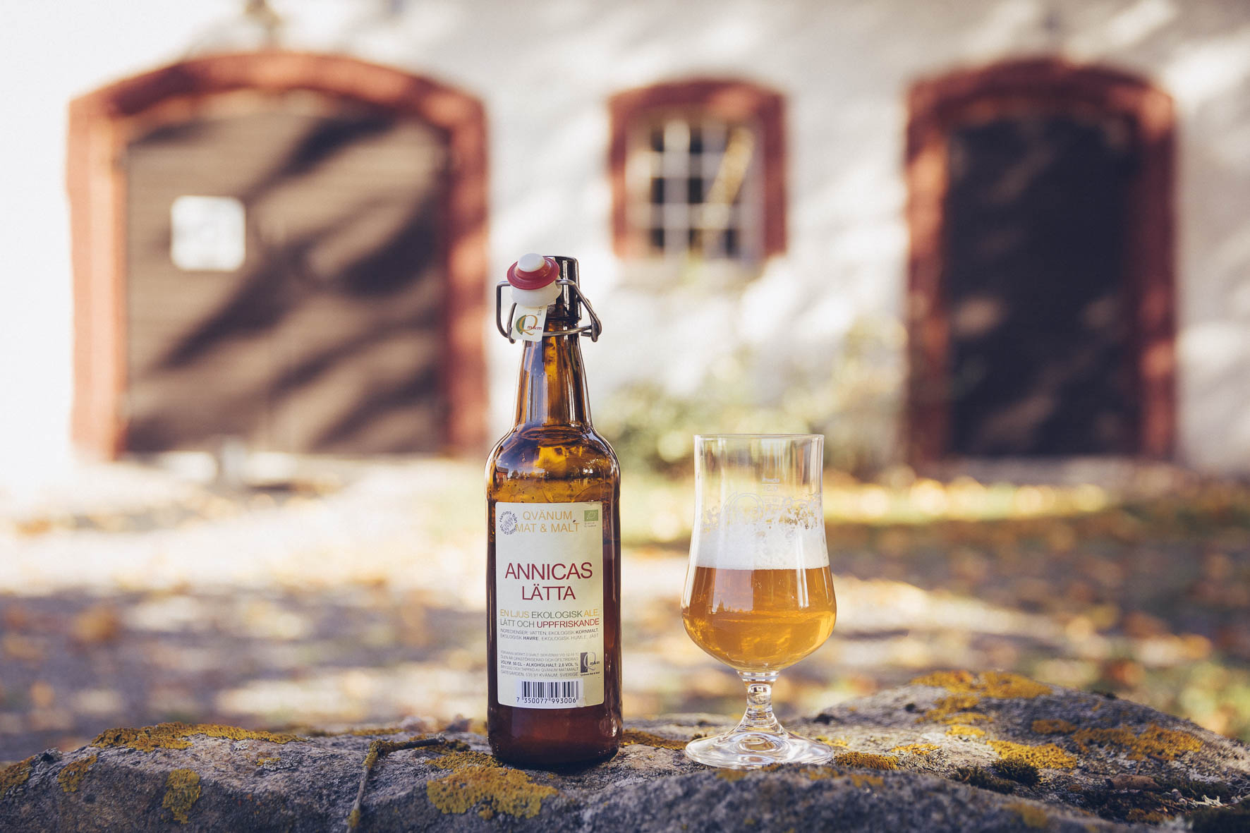 En produktbild på en öl och ett upphällt glas framör en välskött ladugård.