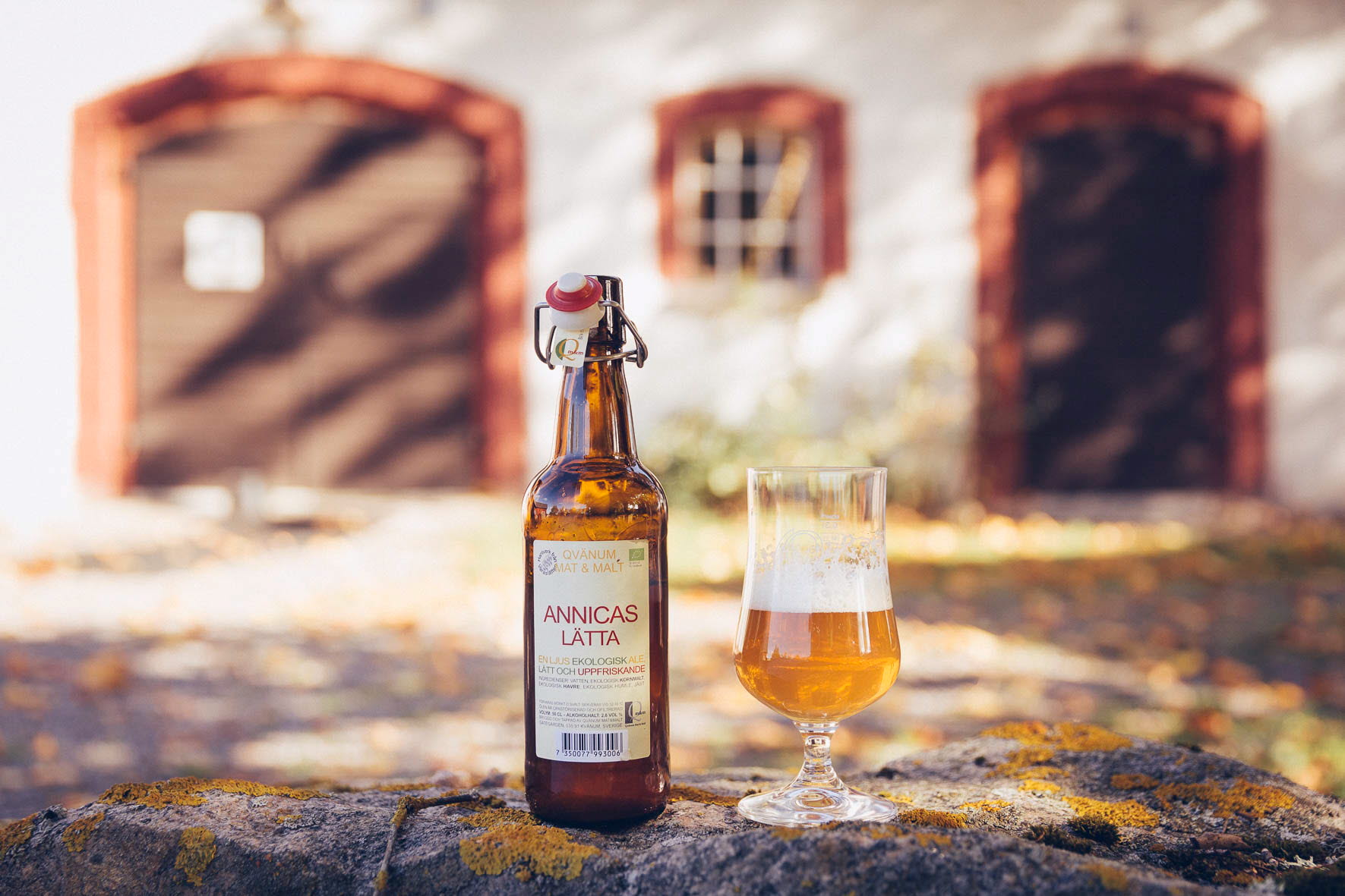 En produktbild på en öl och ett upphällt glas framör en välskött ladugård.