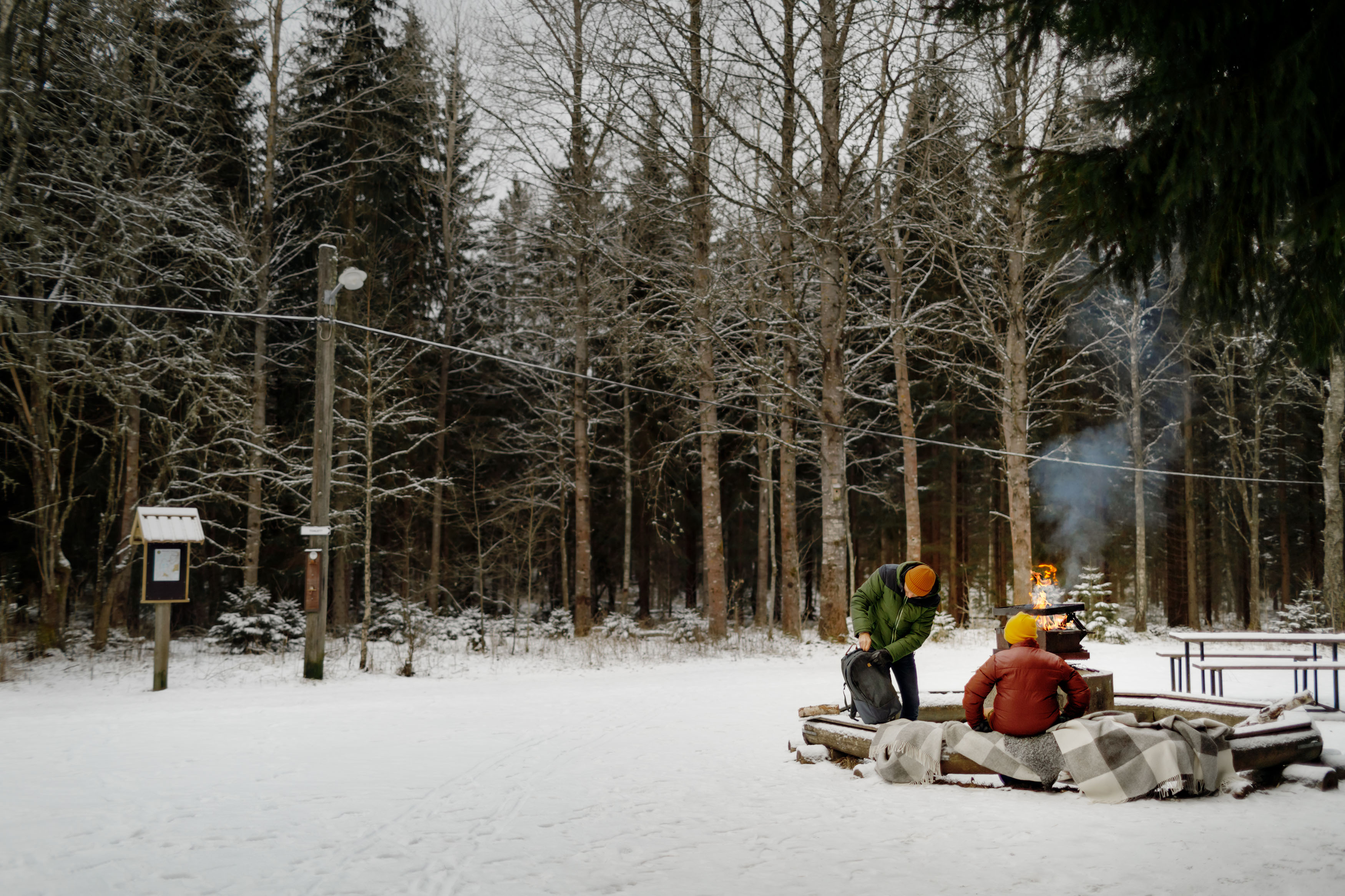 En skog med snö på marken. Vid en eldstad sitter några personer.