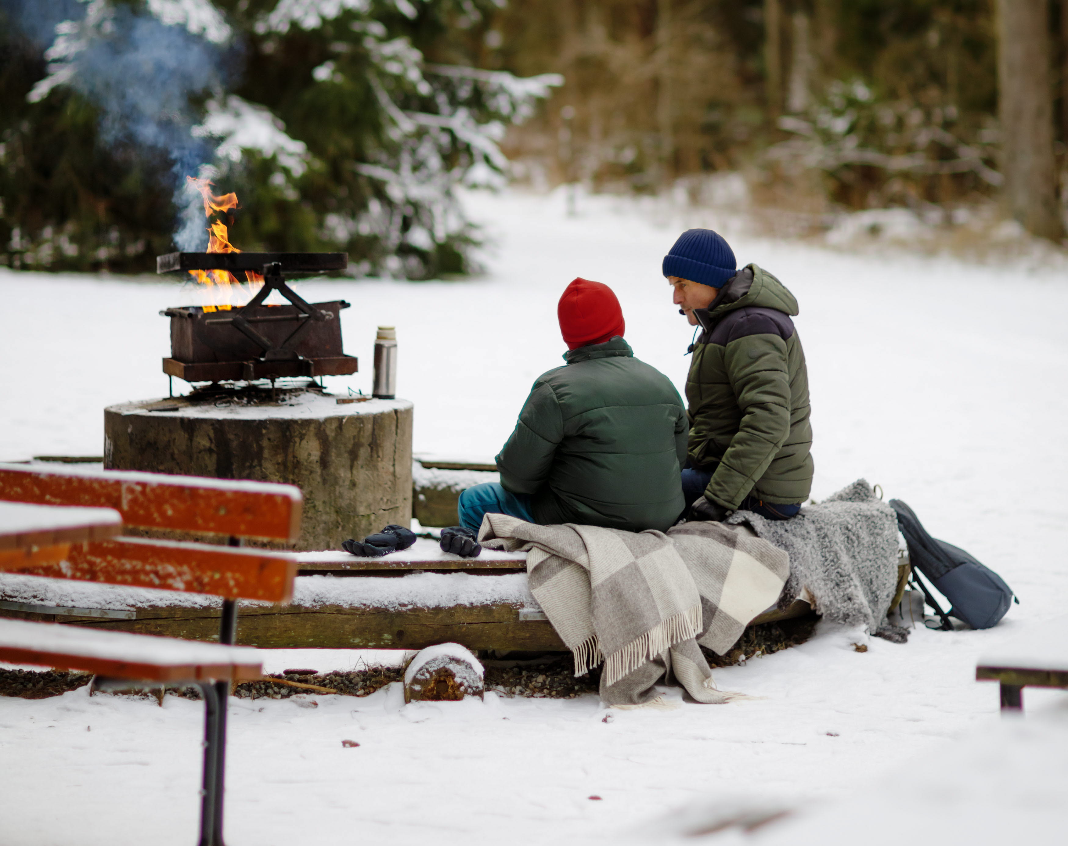 En skog med snö på marken. Vid en eldstad sitter några personer.