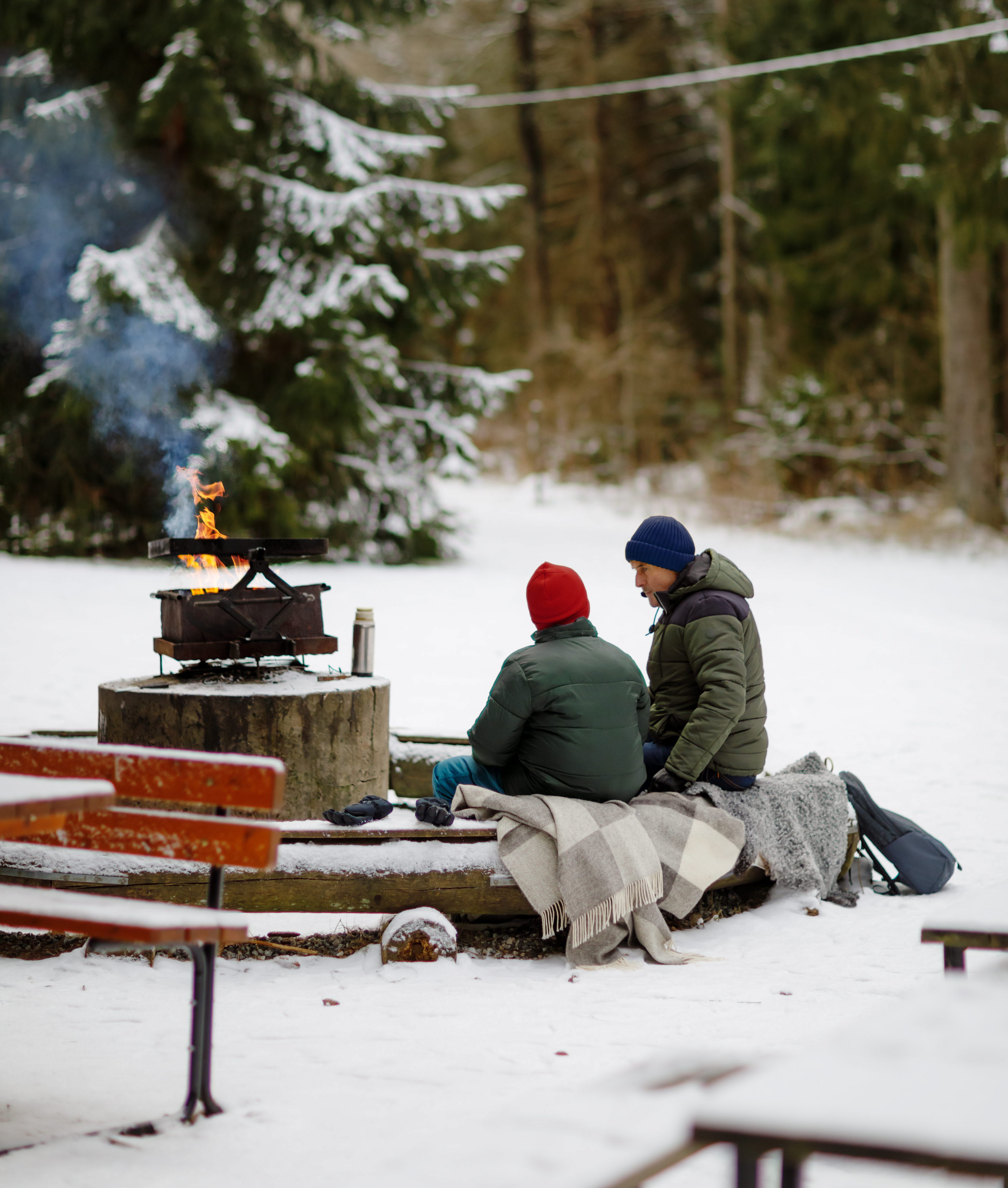 En skog med snö på marken. Vid en eldstad sitter några personer.