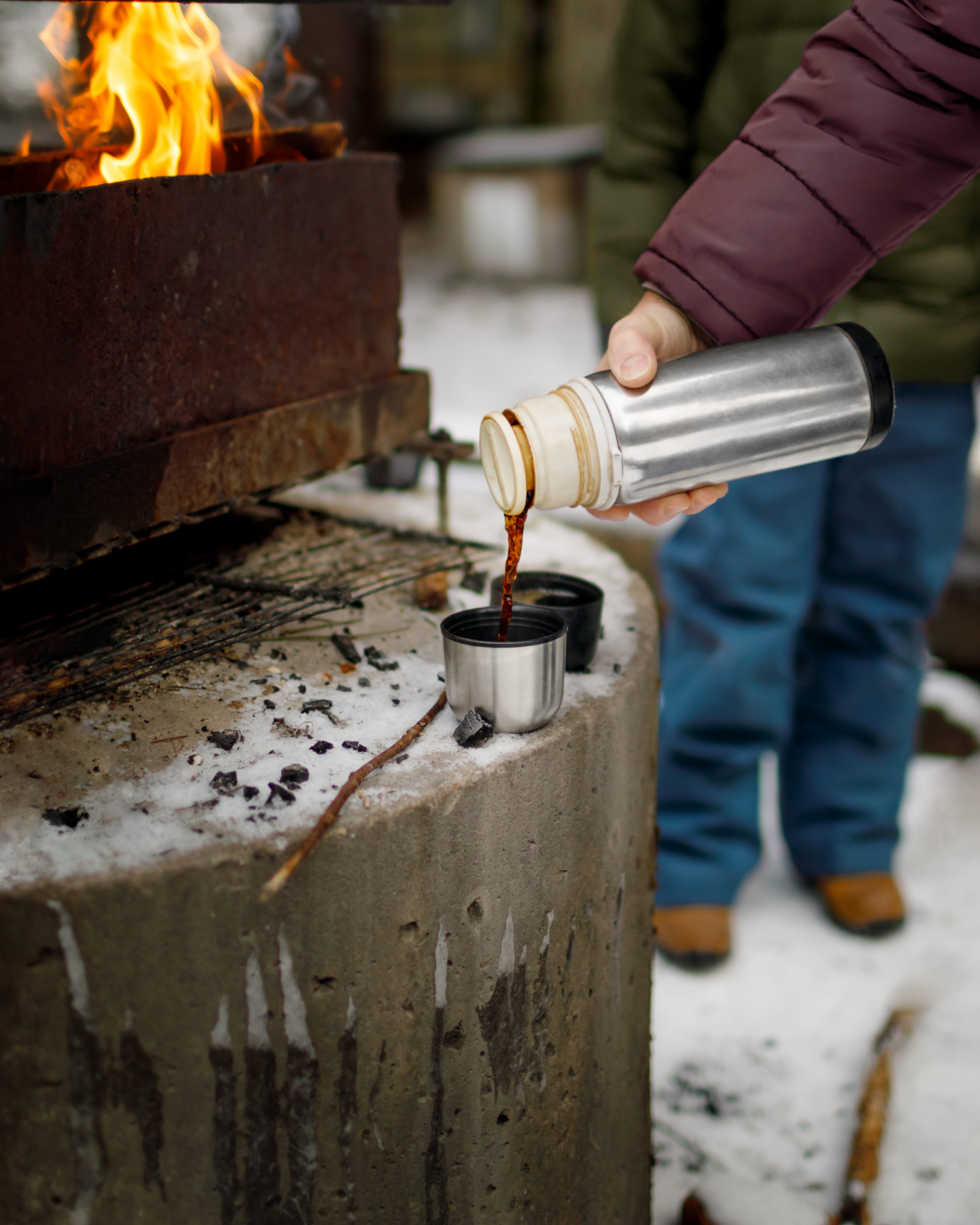En person som håller i en termos och häller upp kaffe. I bakgrunden står ett barn och tittar på. 